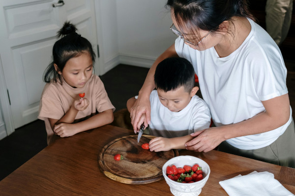 mom showing her little son how to chop a strawberry while the big sister looks on